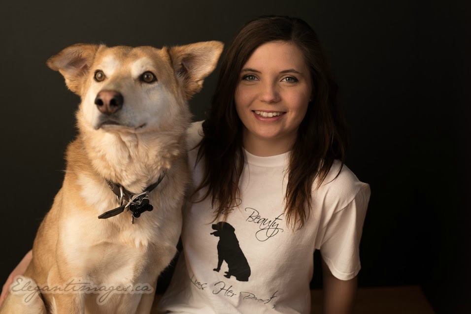 A portrait of a smiling brown haired girl and a tan husky labrador dog. Girl is wearing a white t shirt saying This beauty loves her beast